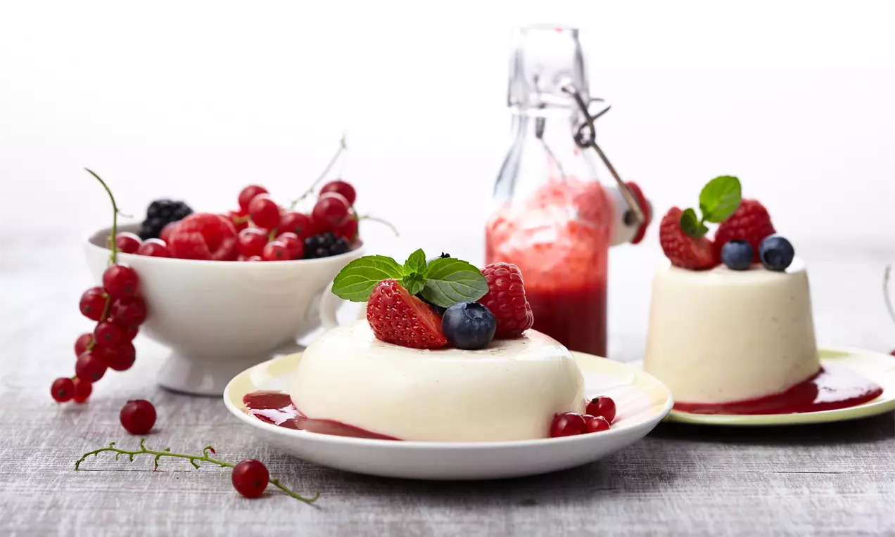 Panna cotta in weißer Glockenform mit glänzender roter Beerensoße, dekoriert mit frischen Beeren und Minzblättern auf einem weißen Teller