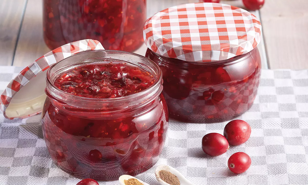 Confettura rosso rubino ricca di frutta con consistenza lucida in barattoli di vetro chiusi da coperchi a quadretti, circondata da mirtilli e spezie