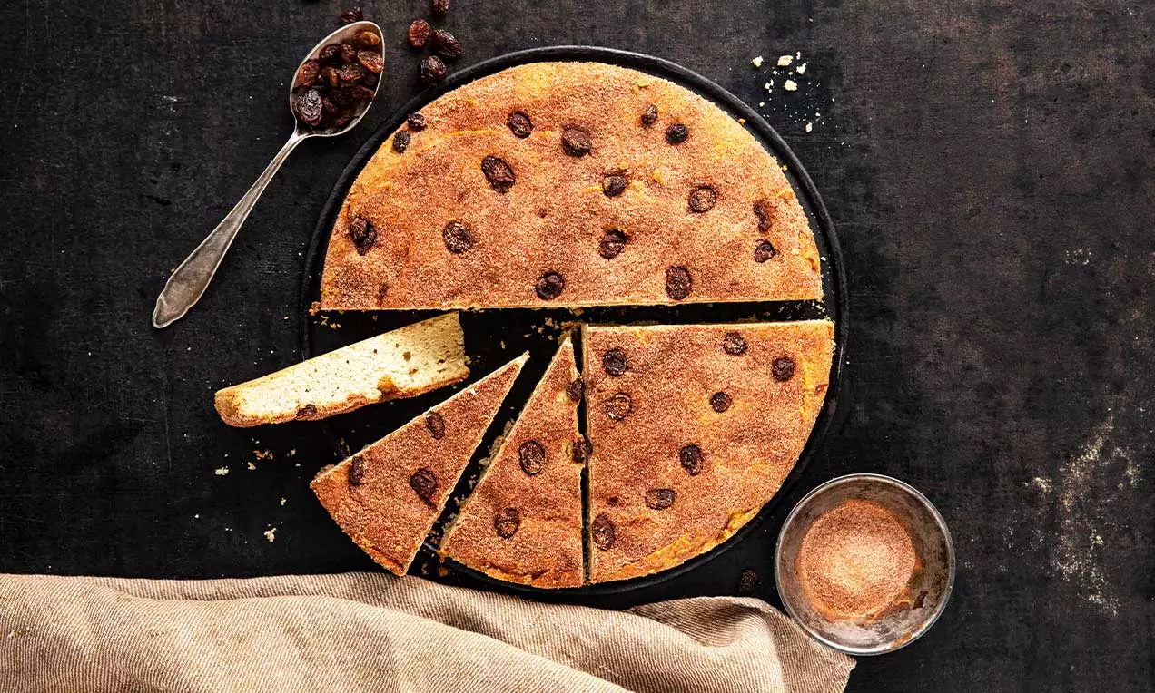 Ein hellbrauner, flacher Kuchen mit goldener Kruste und dunklen Rosinen, in Stücke geschnitten, auf dunklem Untergrund mit Zimt neben einem Löffel