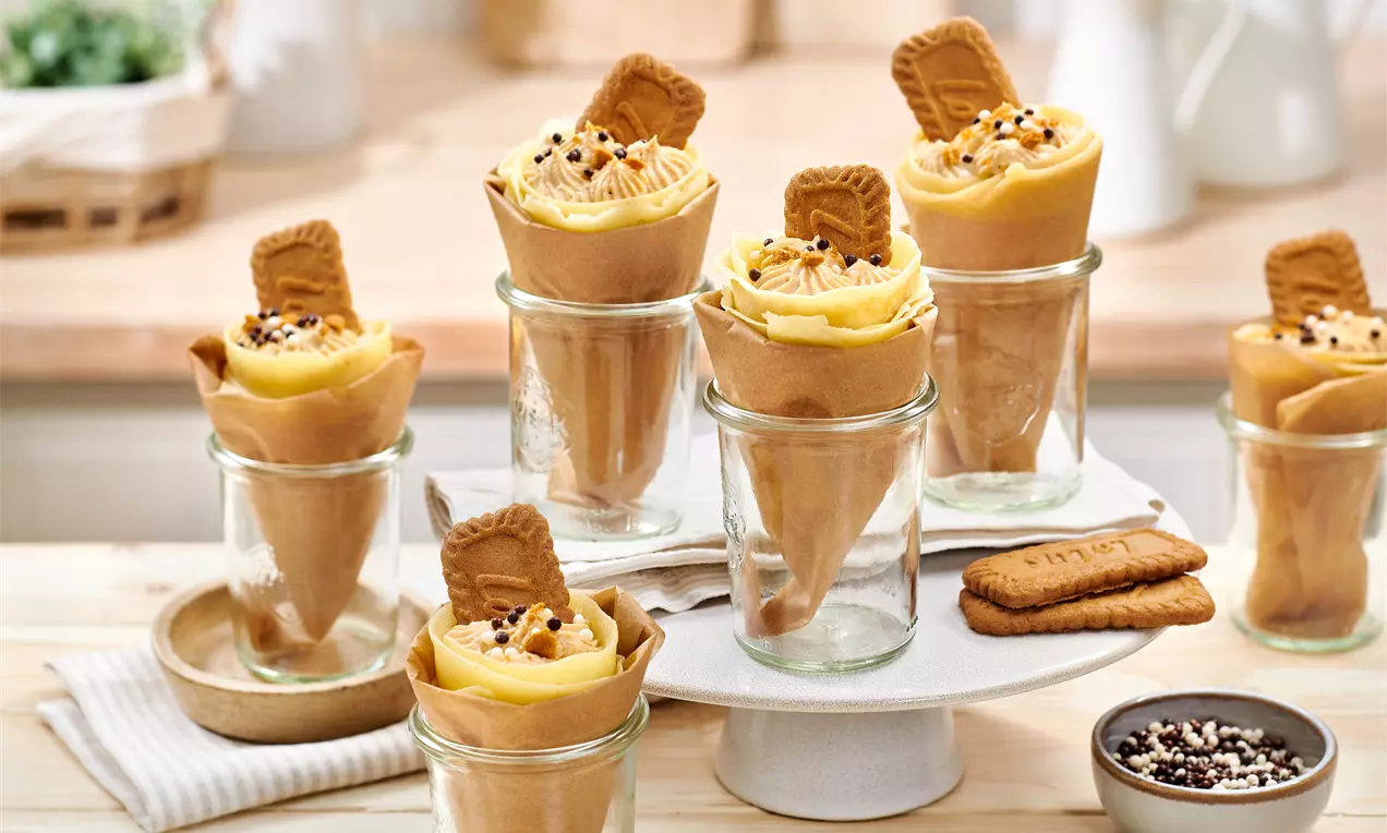 Coni di crepes dorati ripieni di crema Lotus color caramello, decorati con biscotti croccanti e granella su barattoli di vetro e piatto bianco