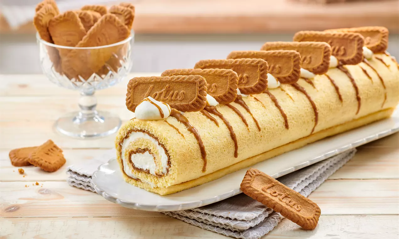 Biscuit roulé doré garni de crème blanche, décoré de biscuits Lotus caramel sur des lignes de sauce Biscoff, présenté sur un plat blanc