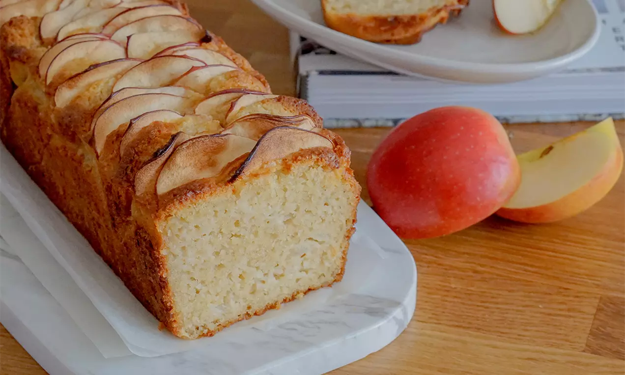 Goldbrauner Kastenkuchen mit luftiger Textur, bedeckt von dünnen Apfelscheiben, auf einer weißen Platte neben frischen Äpfeln und einem Teller