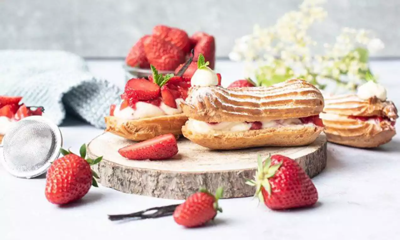 Goldene, längliche Vanille-Eclairs mit geriffelter Oberfläche, gefüllt mit heller Creme und frischen Erdbeerscheiben, auf einer Holzplatte drapiert