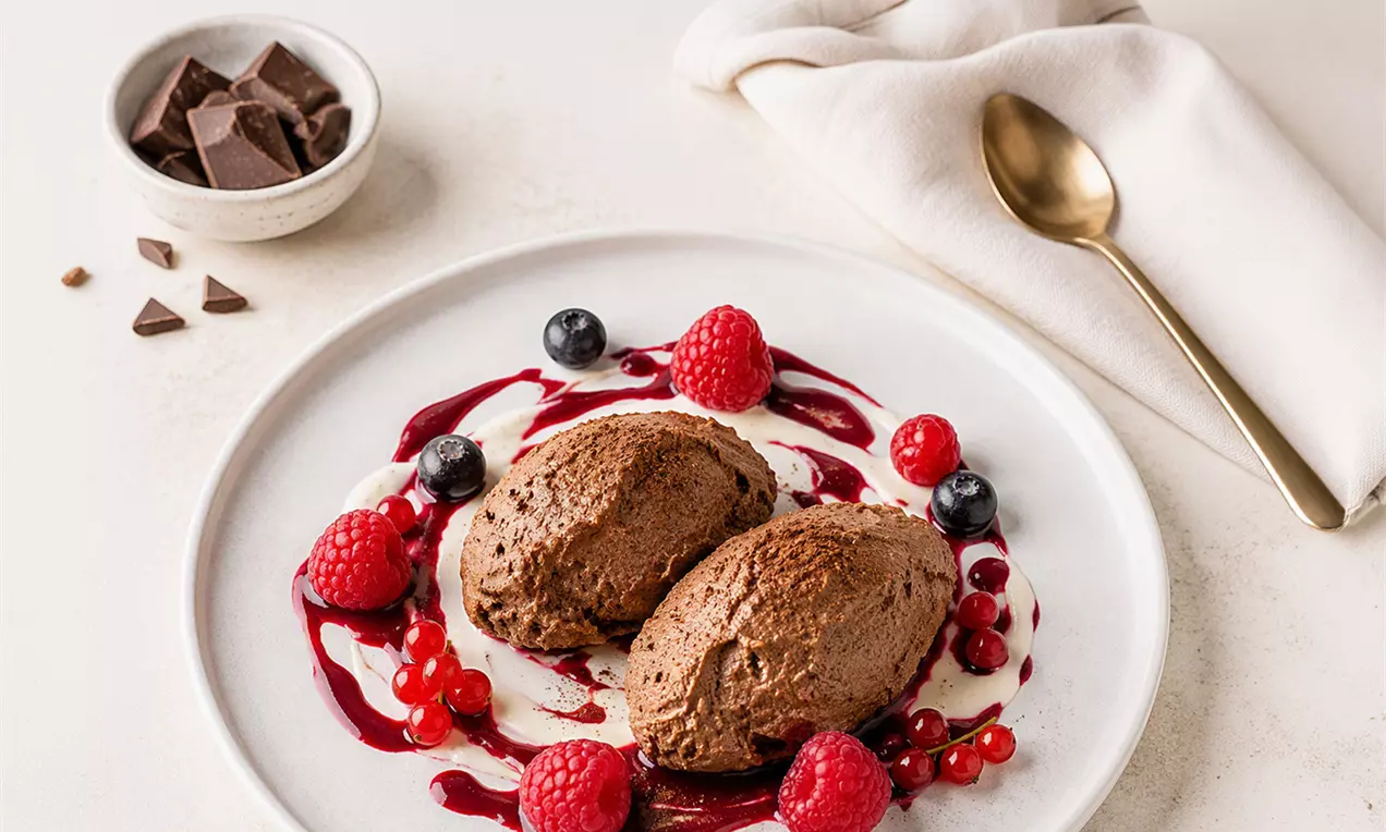 Zwei dunkelbraune Schokomousse-Nocken auf weißem Teller, eingerahmt von Beeren und roter Fruchtsauce, daneben Löffel und Schokostückchen, AI-labeled image