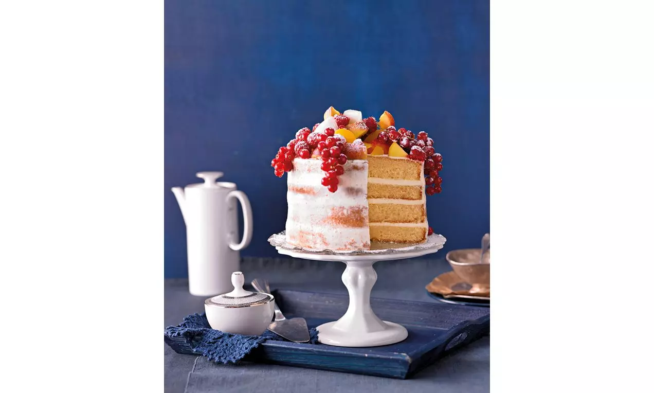 Hohes Schichtdessert mit heller Creme und feinem Biskuit, obenauf frische Früchte wie Johannisbeeren und Pfirsichstücke auf weißer Tortenplatte