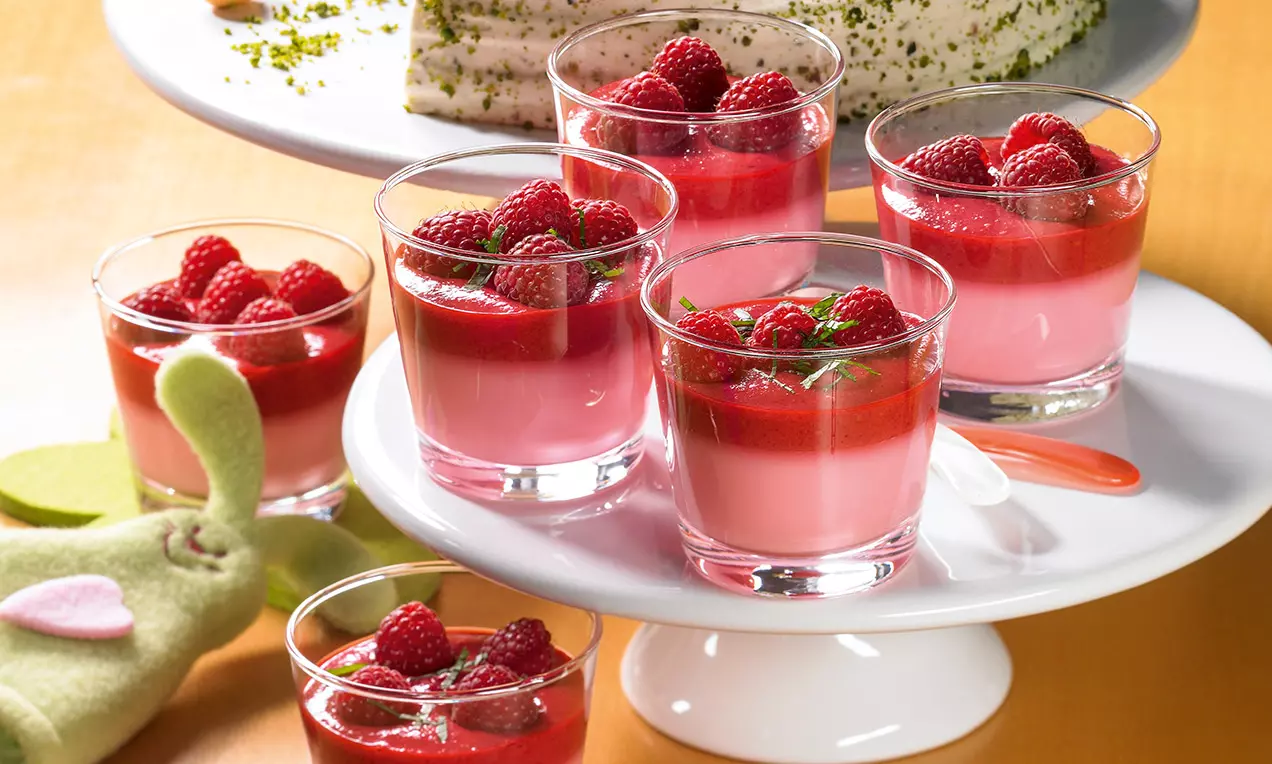 Bicchieri di crema rosa liscia con uno strato di gelatina rossa lucida, guarniti con lamponi freschi e foglioline verdi su un piatto bianco
