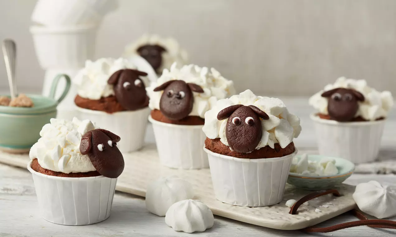 Braune Cupcakes in weißen Förmchen mit weißer Sahnehaube und dunkelbraunen Schäfchen-Gesichtern aus Schokolade auf heller Platte drapiert