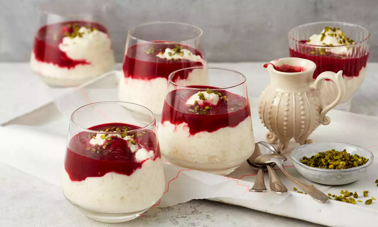 Weiße Gläser mit cremigem Reispudding, leuchtend rotem Fruchtspiegel, Sahnetupfen und gehackten Pistazien auf einem hellen Tisch mit Dekor