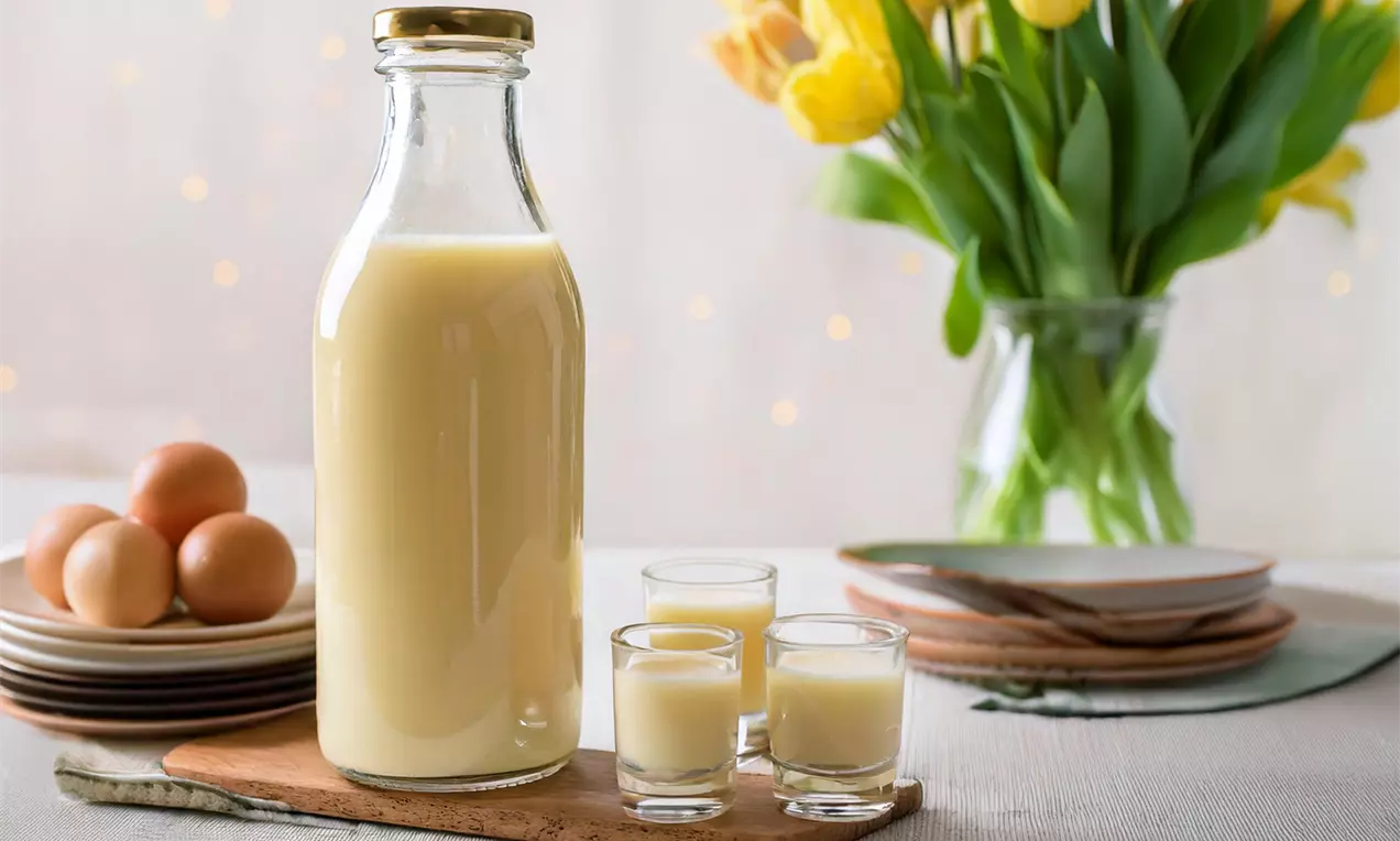 Eine gläserne Flasche mit hellgelbem Eierlikör neben drei gefüllten Shotgläsern auf einem Holzbrett, umgeben von Eiern und gelben Tulpen