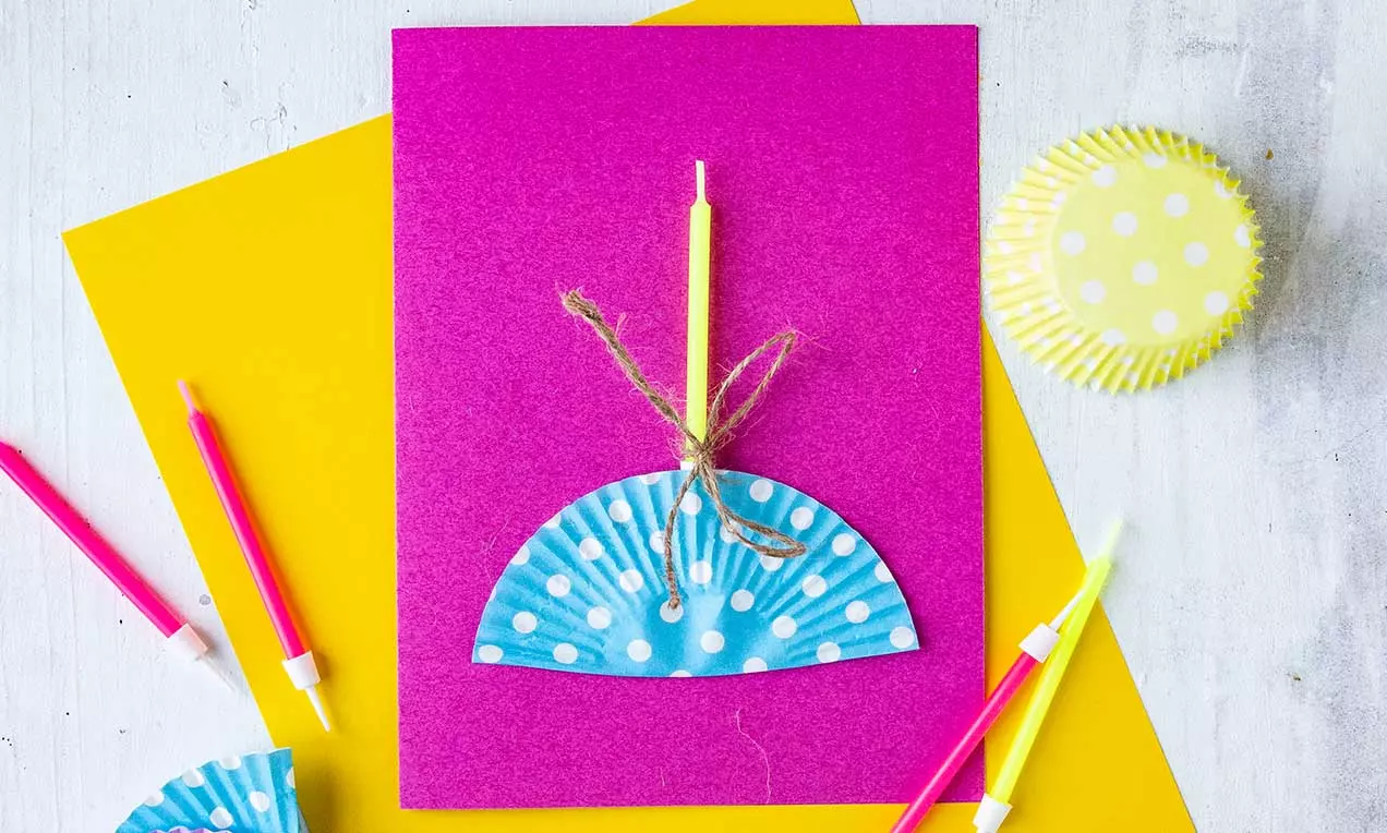 Carte rose vif décorée d’un demi-moule à muffin bleu à pois blancs lié avec une corde, entourée de bougies et d’un moule jaune