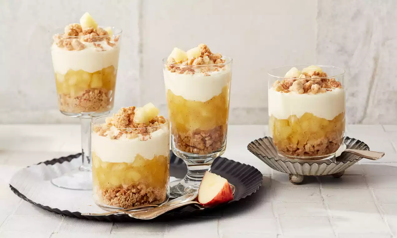 Apfelkuchen im Glas mit goldgelber Apfelkompott-Schicht, heller Creme, knusprigen Streuseln und Apfelstücken auf Tablett mit Löffel und Apfelschnitz