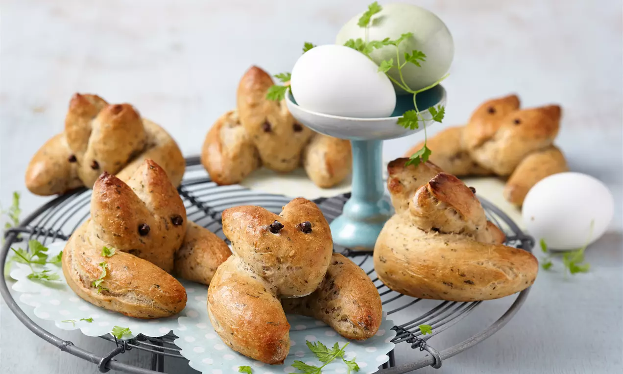 Petits lapins dorés en pâte levée parsemée d'herbes, disposés sur une grille noire avec des œufs blancs et verts en arrière-plan