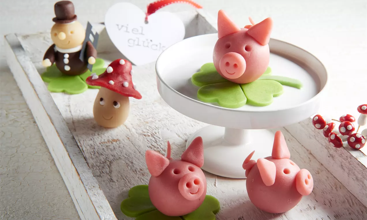 Rosa Marzipan-Säuli mit rundem Körper und kleinen Ohren, platziert auf grünen Kleeblättern aus Marzipan, auf Holztablett dekoriert