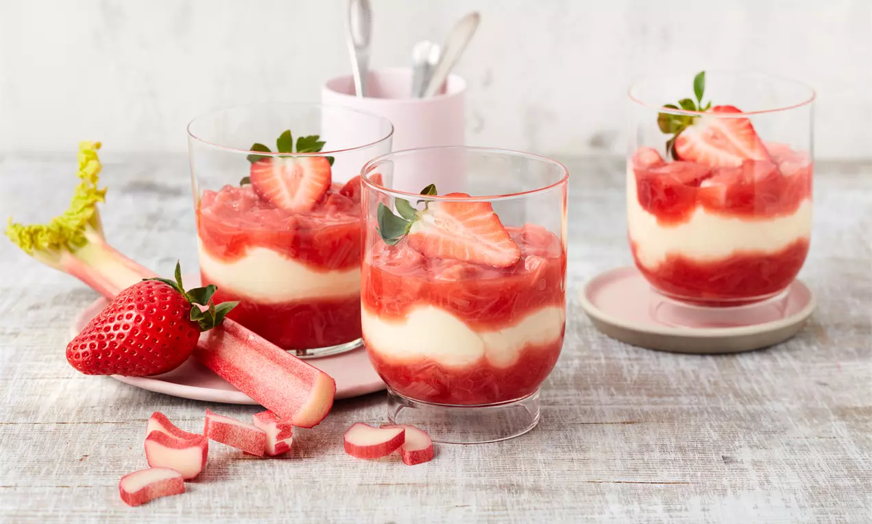 Erdbeer-Rhabarber-Dessert in klaren Gläsern mit weißer Creme, roter Fruchtschicht, frischen Erdbeeren und Rhabarberstücken auf hellem Holz