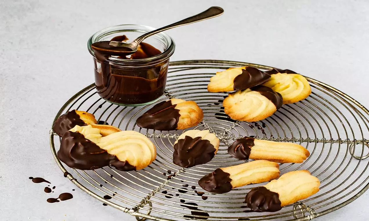 Biscotti ondulati color crema dorato con punte immerse in cioccolato fondente su una griglia con un vasetto di salsa al cioccolato accanto
