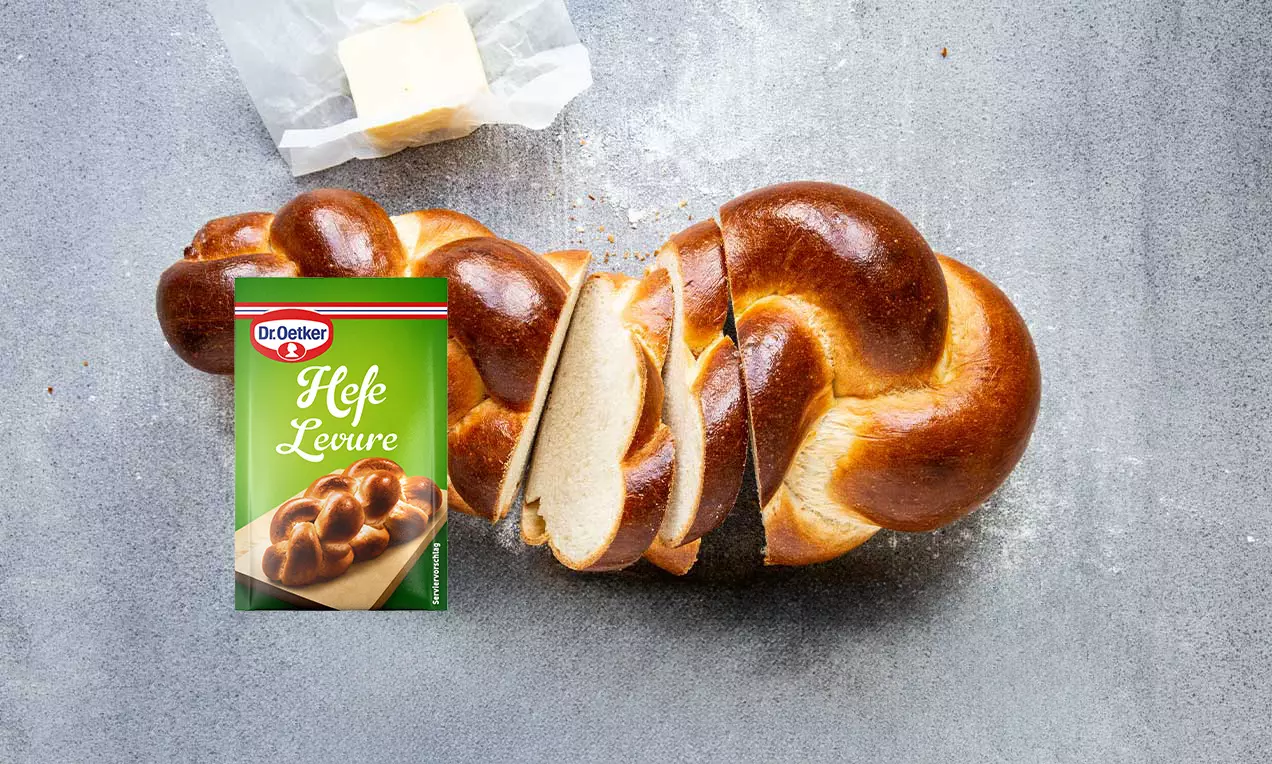 Treccia di pane lucido color caramello con interno soffice e bianco, affettata su una superficie grigia accanto a burro e lievito