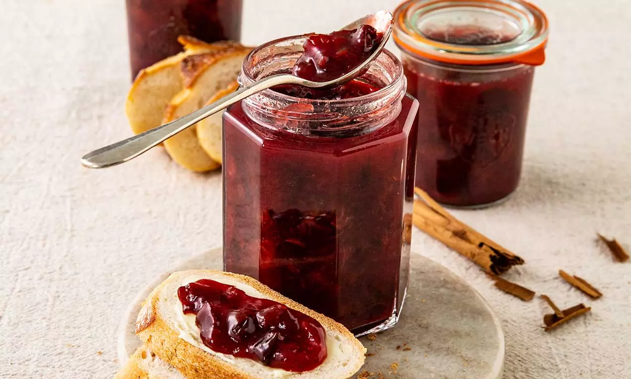 Glas mit dunkelroter Zwetschgen-Zimt-Konfitüre, daneben bestrichene Brotscheiben und Stangen Zimt auf hellem Untergrund