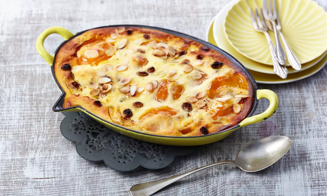 Goldgelber Quark-Gratin mit Aprikosen, Rosinen und Mandelblättchen in ovaler gelber Form auf dunkler Unterlage mit silbernem Löffel