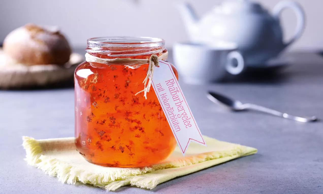 Ein durchsichtiges Einmachglas mit leuchtend orangem Rhabarbergelee und Holunderblüten, dekorativ auf gelber Stoffserviette platziert