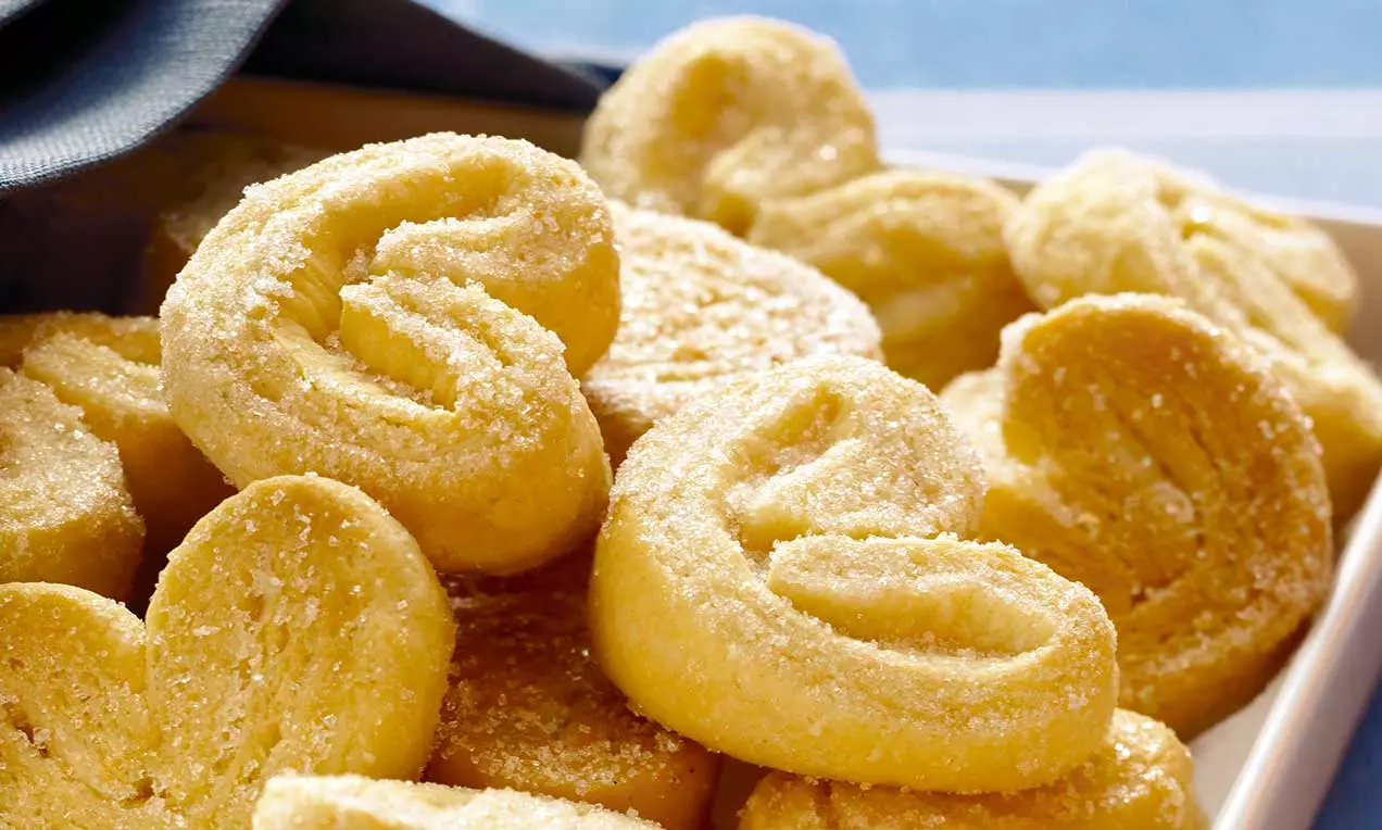 Biscotti dorati a forma di orecchie di maiale con superficie croccante ricoperta di zucchero, disposti su un piatto chiaro tra ombre morbide
