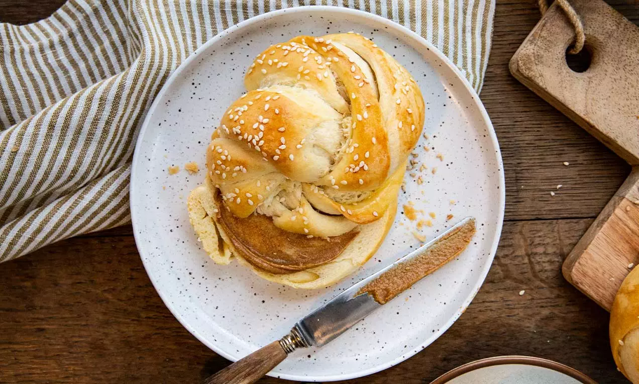 Petit pain turc doré et tressé, saupoudré de graines de sésame sur une assiette blanche, accompagné d’un couteau en bois sur une table rustique