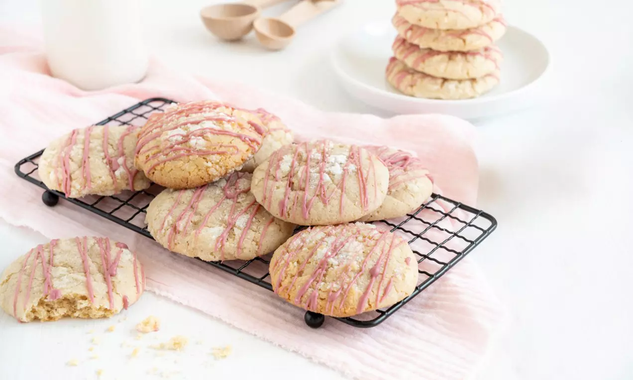 Vanille Cookies mit zarter rosa Glasur auf einem schwarzen Gitterrost, umgeben von hellem Stoff, mit Krümeln und Messlöffeln im Hintergrund