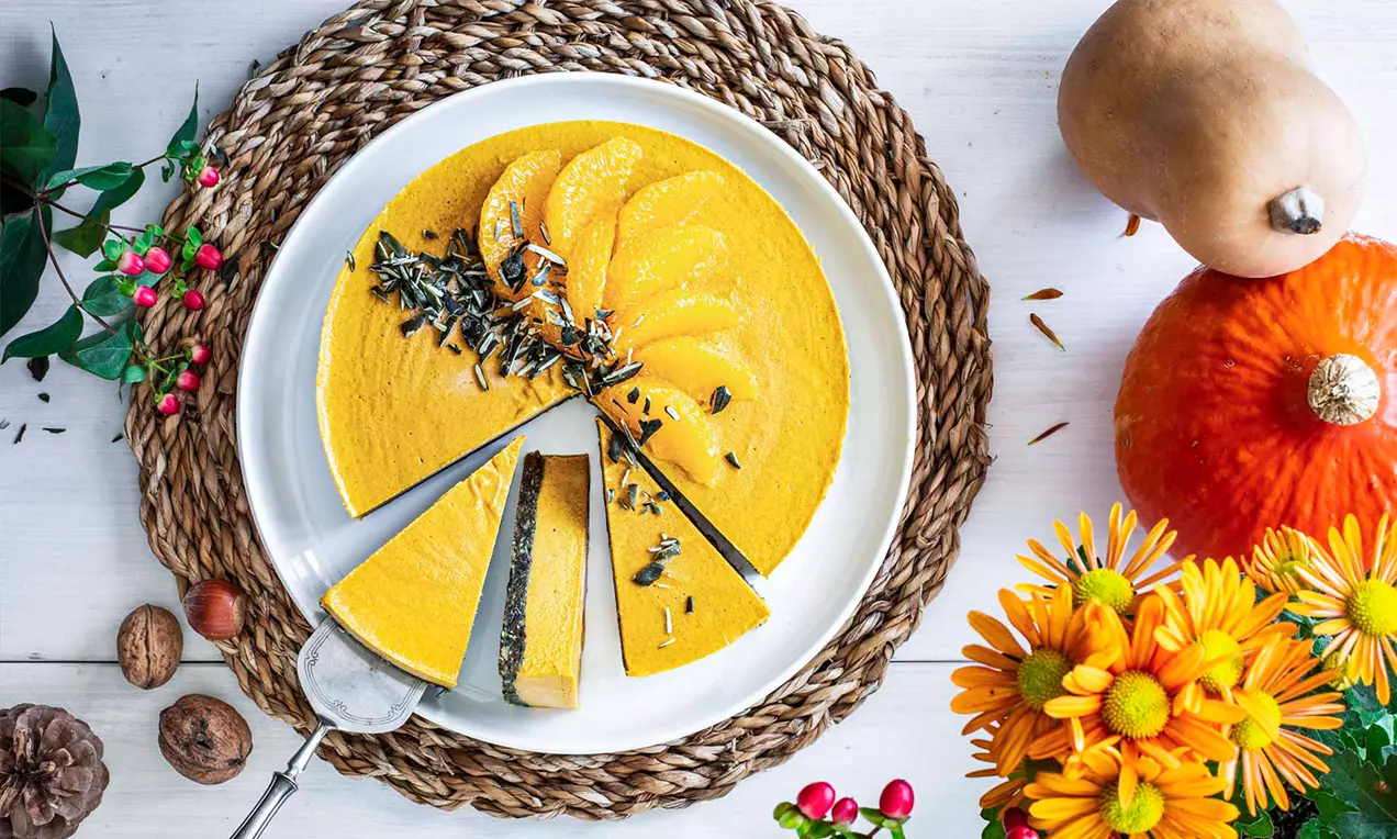 Cheesecake senza cottura alla zucca su base croccante, dal colore giallo brillante con fettine di zucca e semi disposti a ventaglio su un piatto bianco