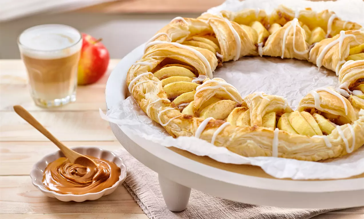 Torta di mele su base di sfoglia dorata intrecciata, con fettine di mela giallo tenue e fili di glassa bianca, su un piatto tondo bianco