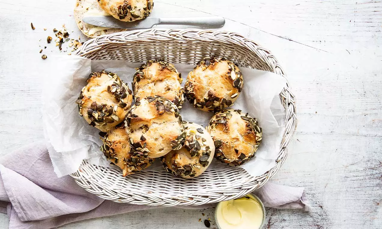 Goldbraune Marroni-Brötli mit knuspriger Kruste und Kürbiskernen in einem geflochtenen Korb auf weißem Papier mit Butter und Stofftuch