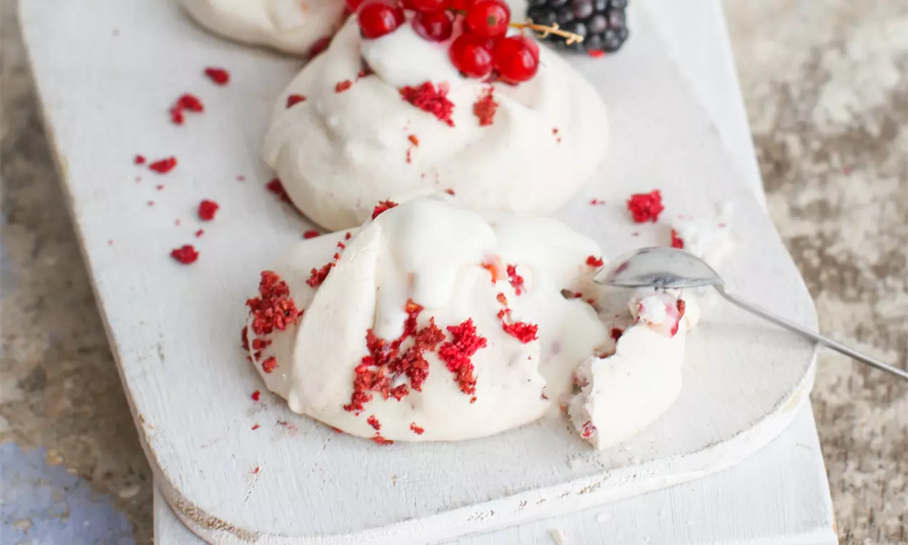 Weiße Mini-Pavlovas mit knuspriger Kruste, cremiger Füllung und roten Beeren auf einem rustikalen Holzbrett angerichtet