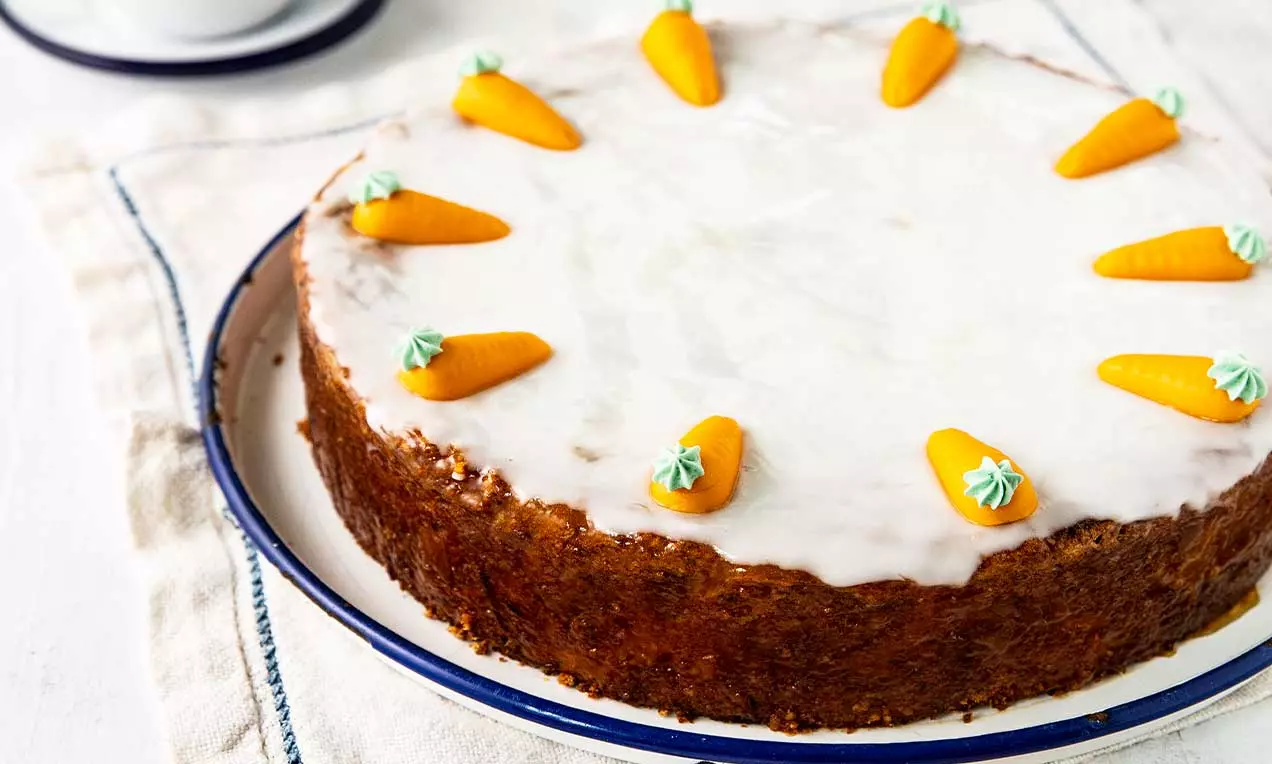 Torta rotonda con glassa bianca lucida, bordi dorati e decorazioni di carotine arancioni in pasta di mandorle con ciuffi verdi