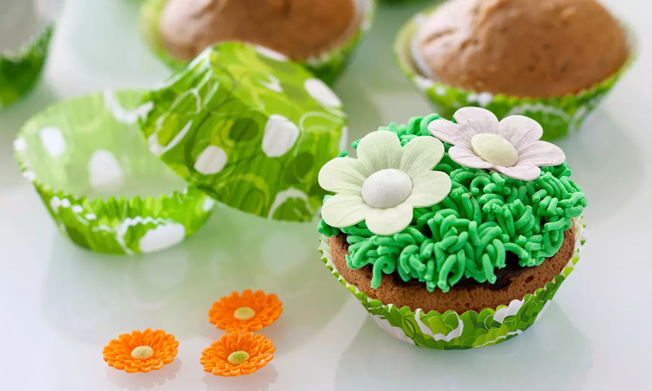 Cupcake garni de crème verte en forme d'herbe, décoré de fleurs en sucre blanc et rose, posé dans une caissette verte à motifs blancs