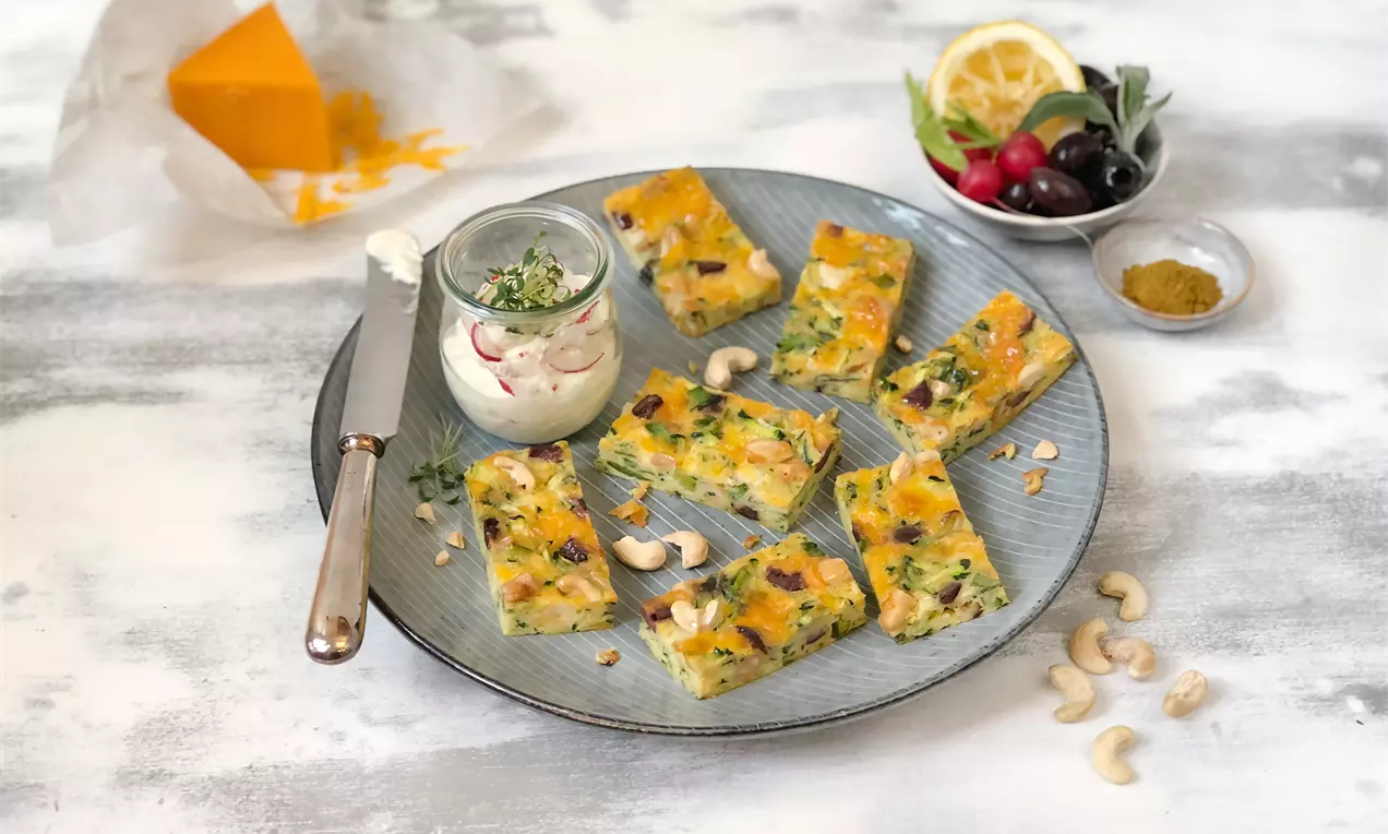 Goldbraune, rechteckige Zucchinikuchenstücke mit Kräutern und Cashewkernen auf einem grauen Teller, dazu Dip in Gläschen und dekorative Beilagen