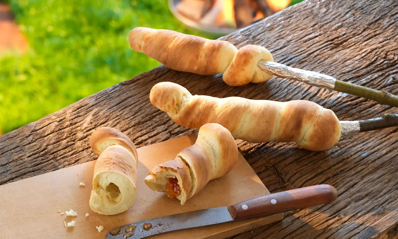 Pâtons dorés et torsadés enroulés autour de bâtons de cuisson sur une planche en bois avec un couteau et un feu crépitant en arrière-plan