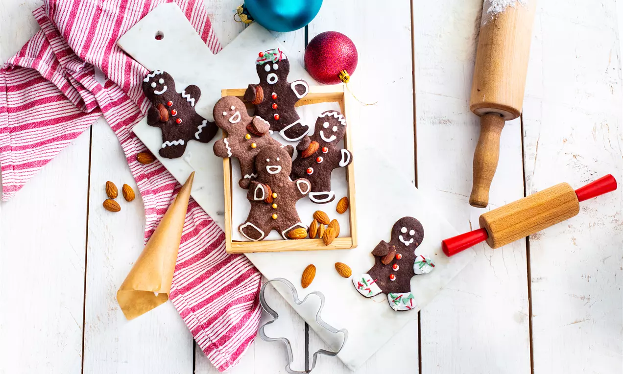 Lebkuchenmännchen aus dunkelbraunem Teig mit bunter Zuckerdekoration auf weißer Platte, umgeben von Mandeln, Weihnachtskugeln und Backzubehör