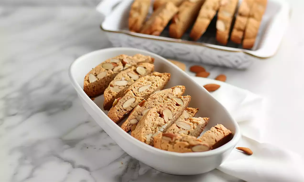 Goldbraune Cantuccini mit knuspriger Textur und sichtbaren Mandeln in einer weißen Schale auf Marmoruntergrund mit lockerem Stoff drapiert
