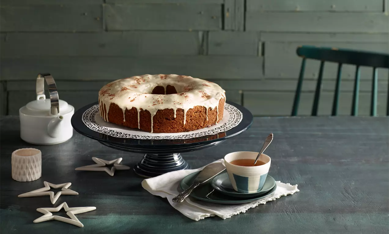 Kranzförmiger Gewürzkuchen mit glänzendem weißem Zuckerguss auf dunkler Tortenplatte, daneben Teetasse und Dekoration mit Sternen
