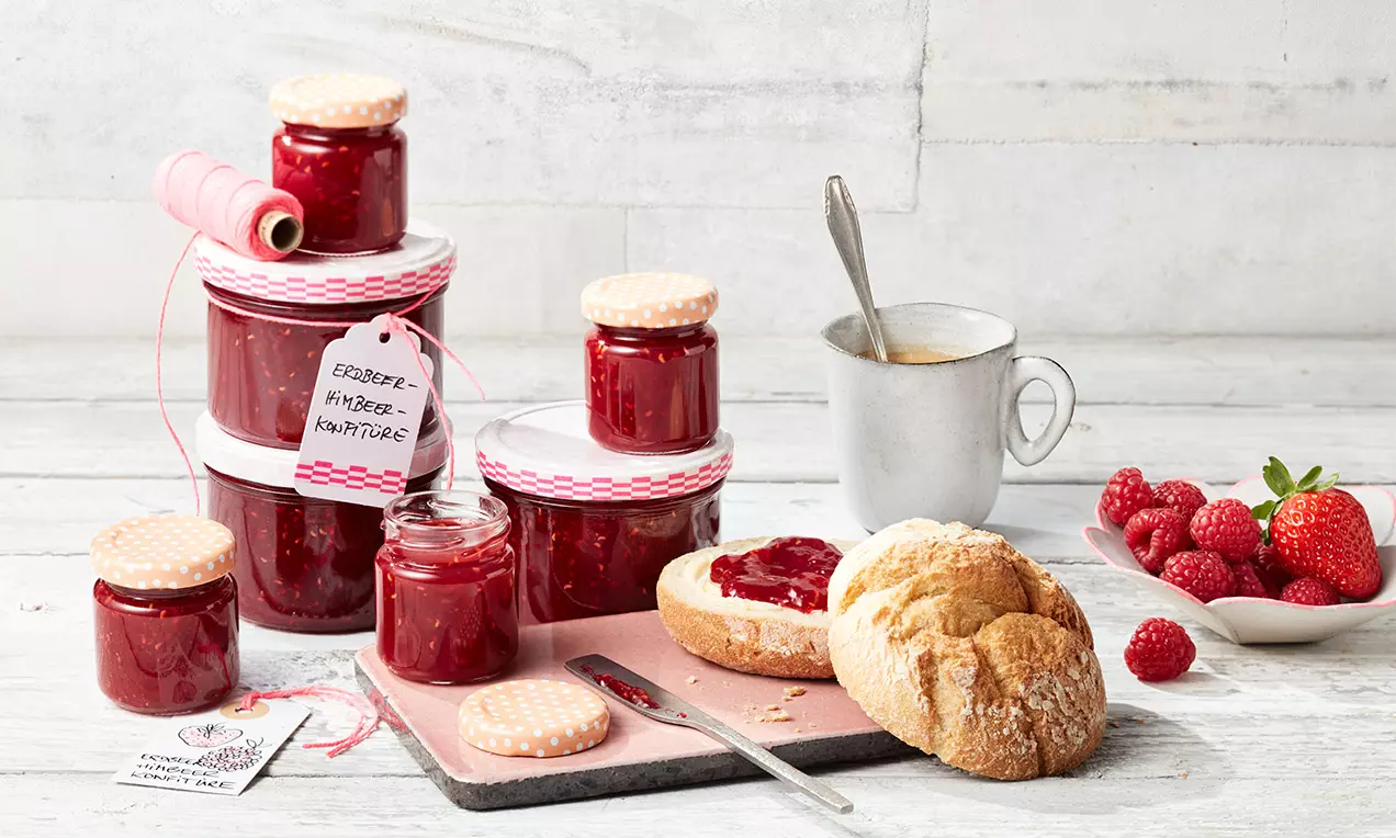 Gläser mit tiefroter Erdbeer-Himbeer-Konfitüre, beigefarbene Deckel mit rotem Muster, daneben ein aufgeschnittenes Brötchen mit Konfitüre