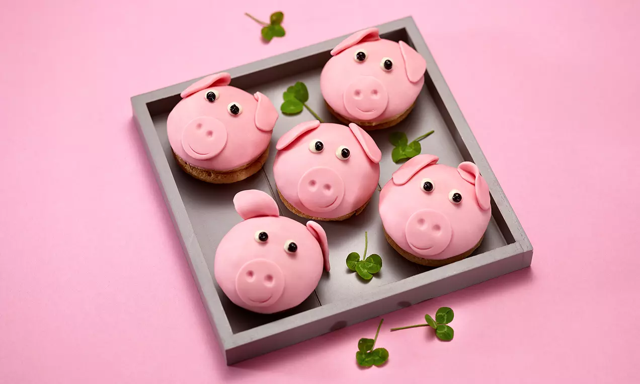 Cupcake decorati con copertura rosa a forma di testa di maialino con occhi neri, orecchie scolpite e trifogli verdi su un vassoio quadrato grigio