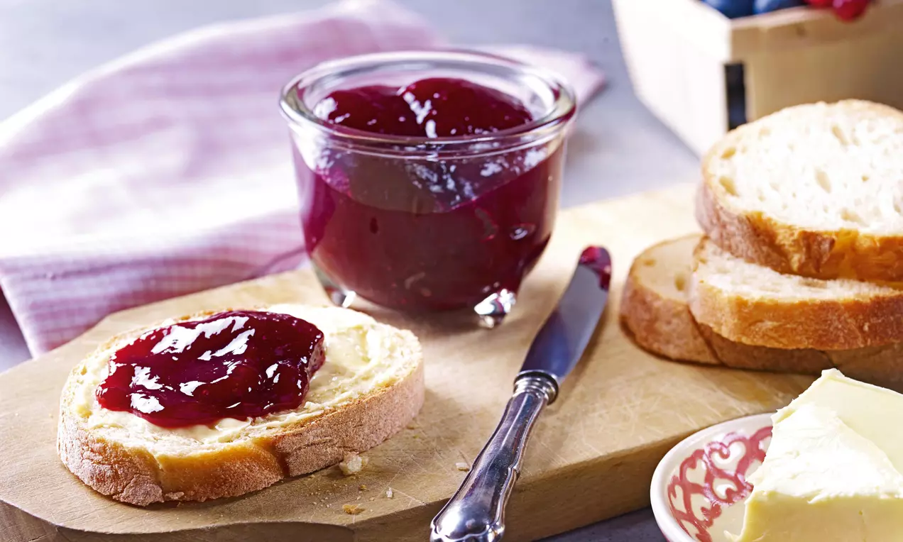 Glas mit glänzend dunkelroter Heidelbeer-Johannisbeer-Konfitüre auf Holzbrett, daneben frisches Brot, Butter und Beerenkorb im Hintergrund