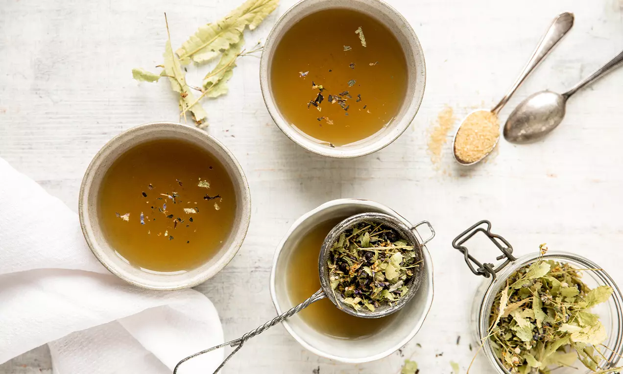 Trois tasses d’infusion ambrée parsemée de fines herbes, avec des feuilles séchées dans un infuseur en métal et un pot en verre nearby