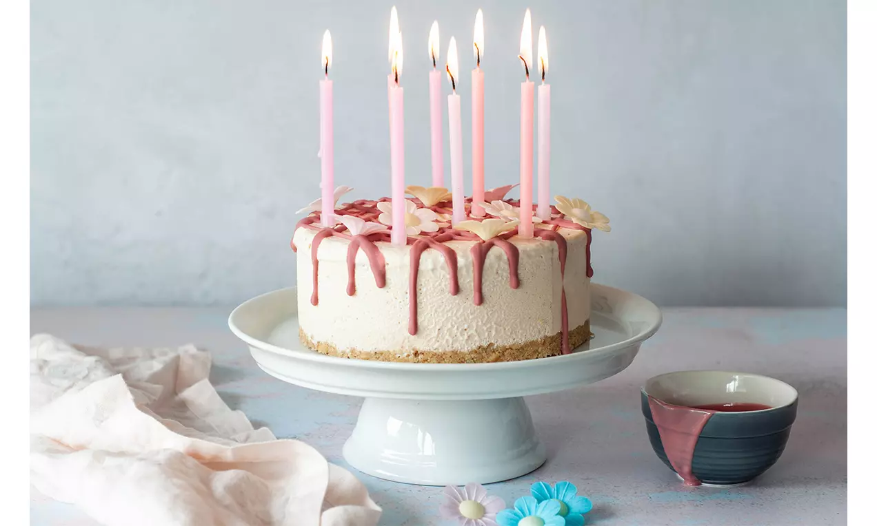Glacé-Torte mit rosa Creme-Tropfen, weißem Überzug, hellem Keksboden, pinken Kerzen und Blumen auf einer weißen Tortenplatte drapiert