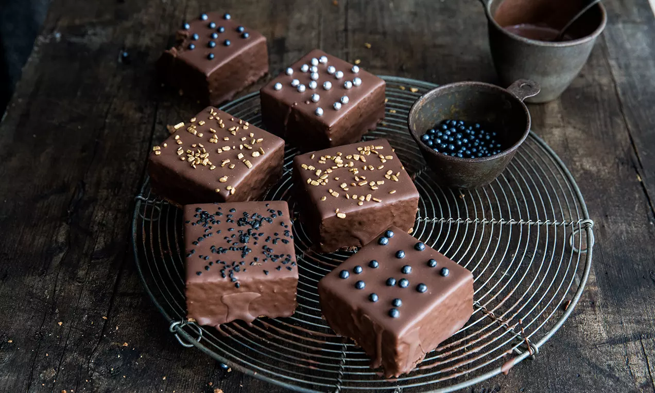 Cubetti di torta rettangolari ricoperti di cioccolato lucido, decorati con granella colorata e disposti su una griglia metallica