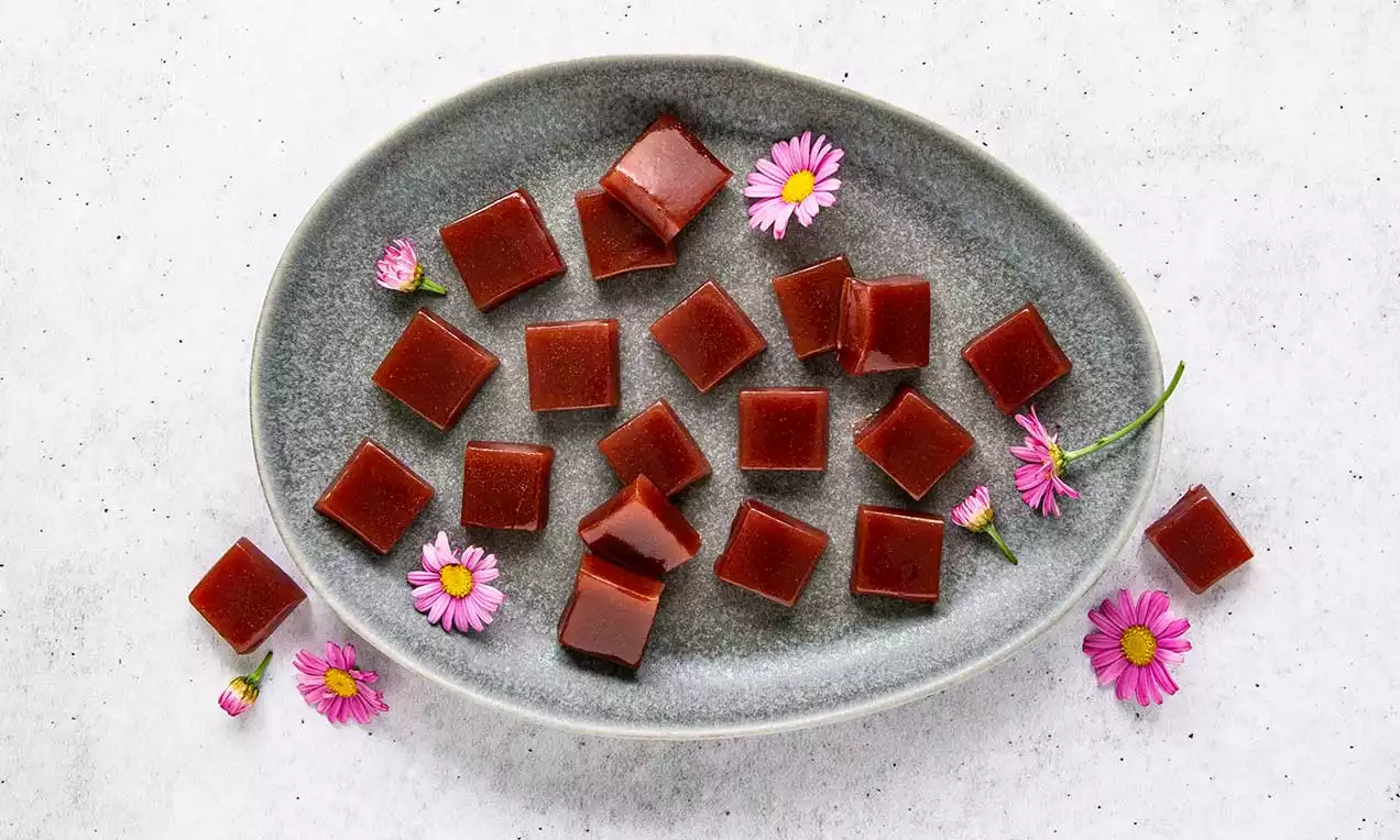 Kleine glänzend rote Würfel mit glatter Oberfläche und karamelligem Glanz, dekoriert mit pinken Blüten auf einem grauen Teller