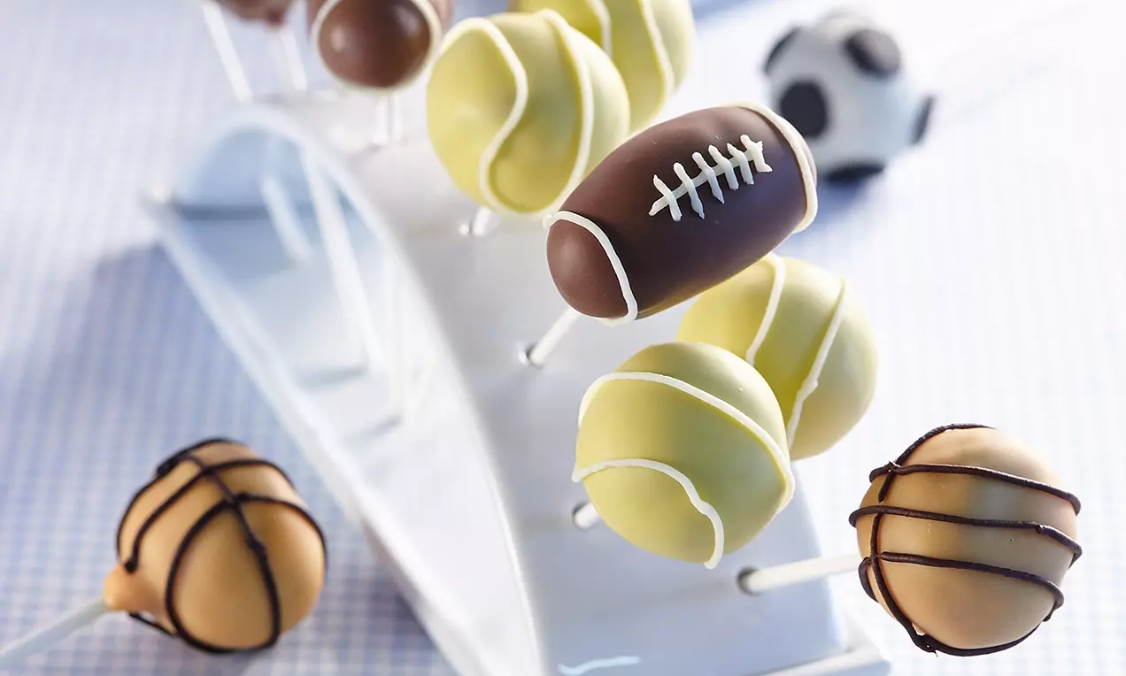 Sucettes cake pop en forme de ballons sportifs colorés, décorées de glaçages lisses et brillants, présentées sur un présentoir blanc