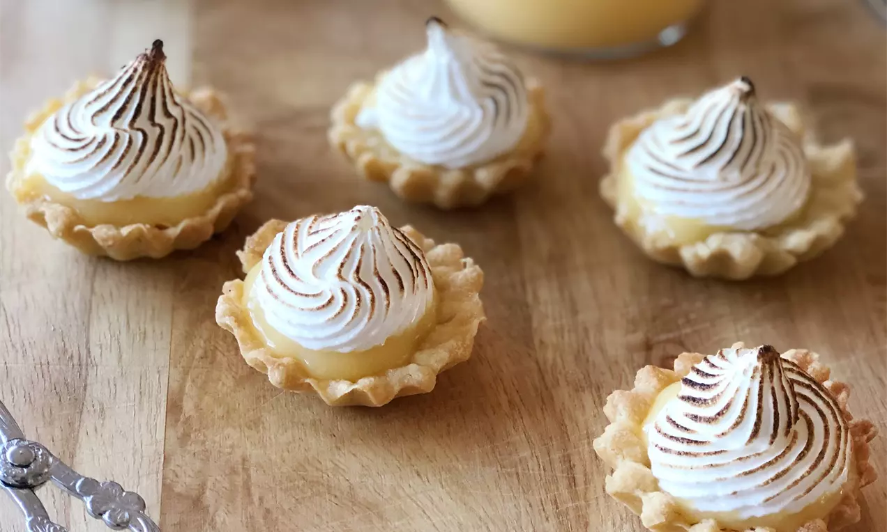 Kleine Törtchen mit goldbraun gebackener Kruste, spiralförmigem Baiserhäubchen und Glasbehälter mit gelber Creme auf Holztisch