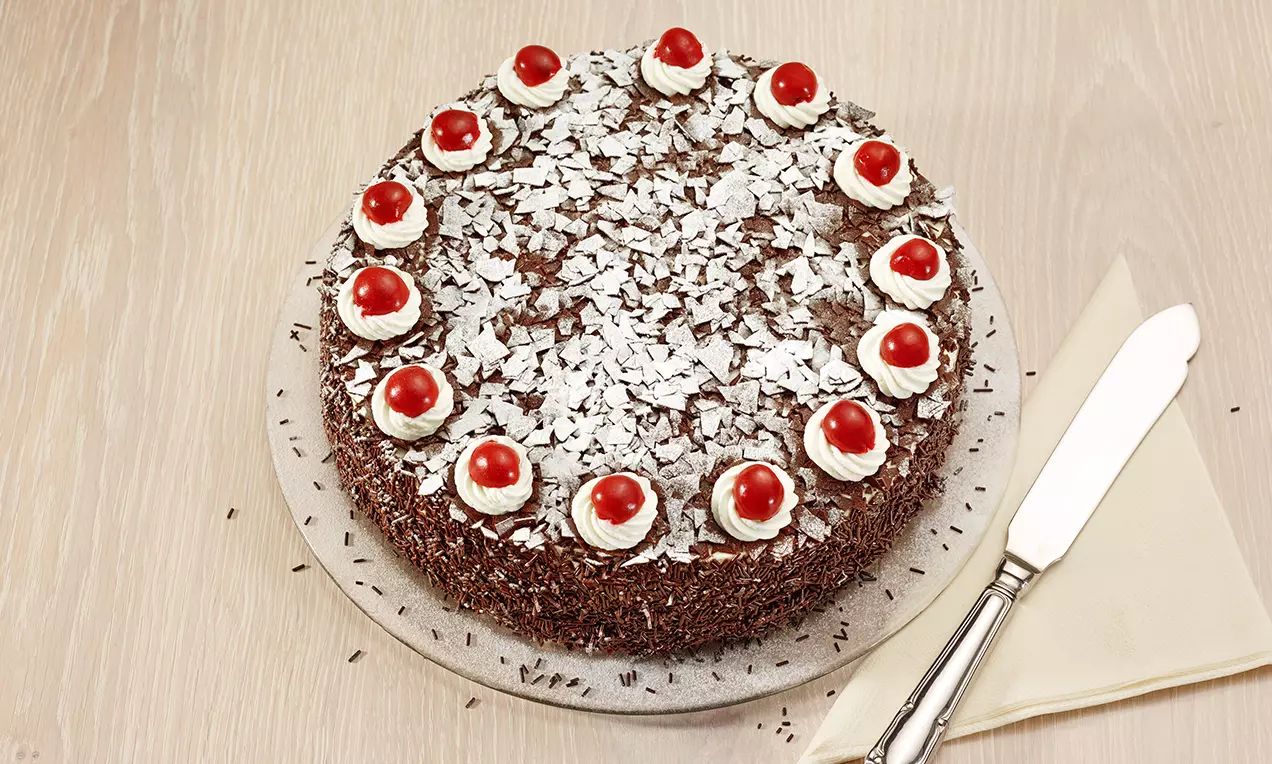 Forêt-Noire ronde avec base chocolatée, décorée de crème blanche et cerises rouges surmontées de copeaux de chocolat argentés sur un plateau