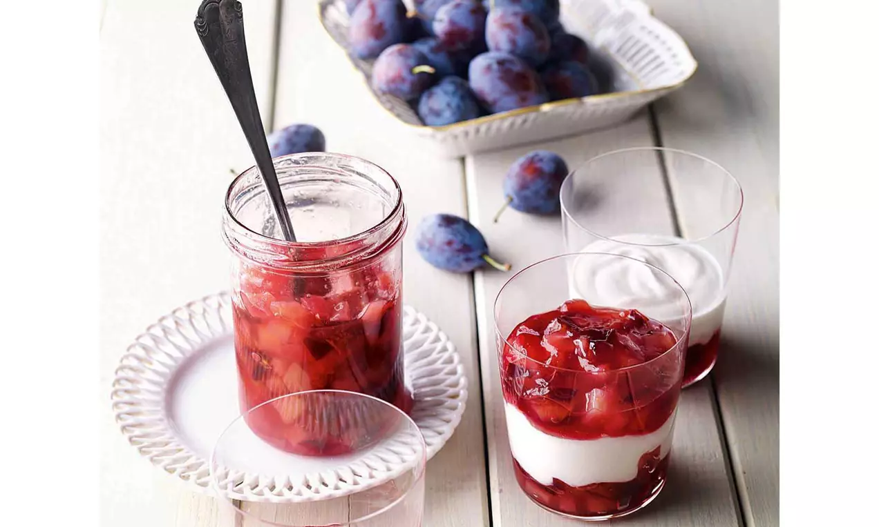 Gerötetes Birnen-Zwetschgen-Kompott mit weichen Fruchtstücken in einem Glas auf einem weißen Teller neben Dessertgläsern und frischen Zwetschgen