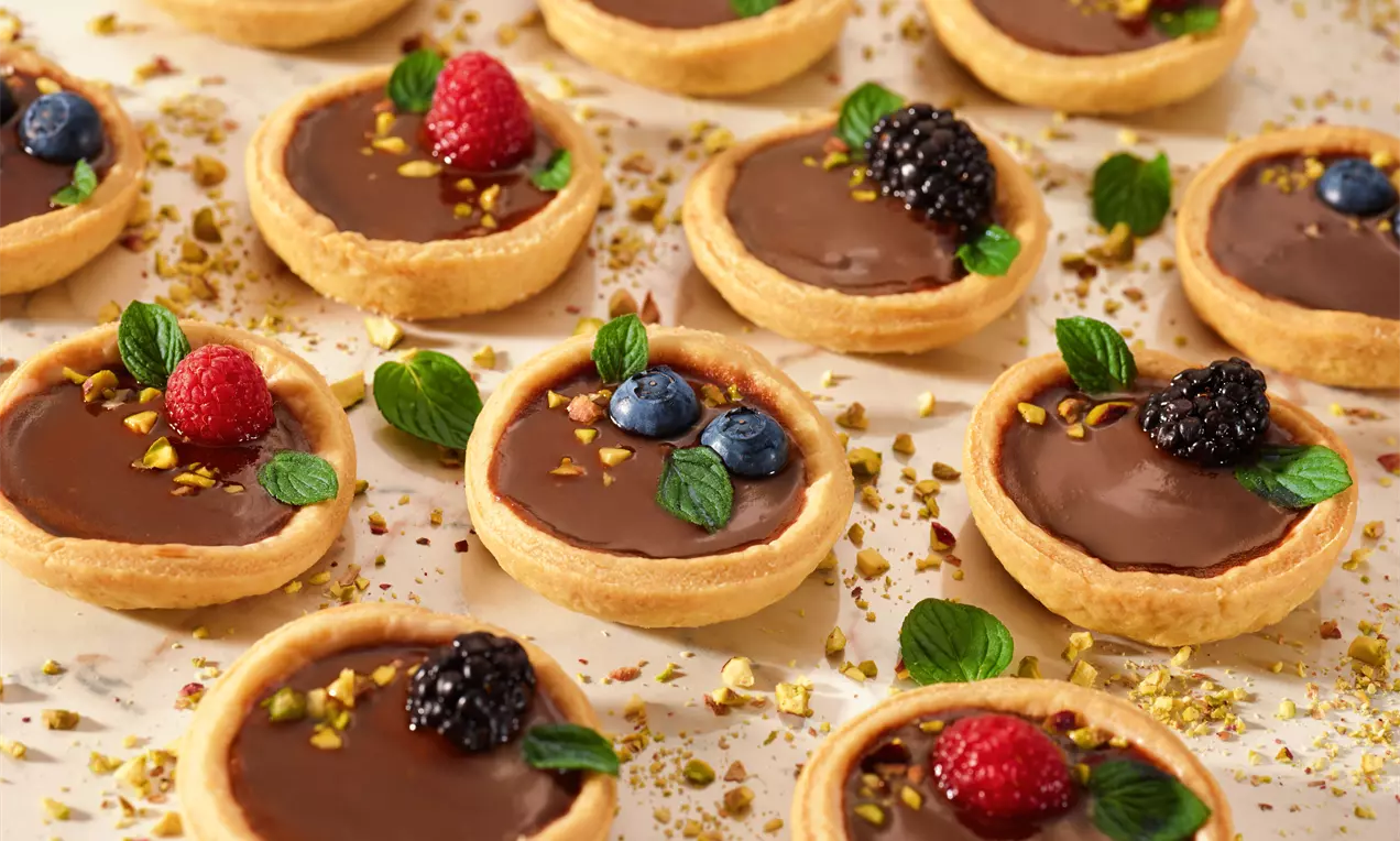 Tortine dorate di pasta frolla tonde con ripieno lucido al cioccolato, guarnite con frutti di bosco freschi, foglie di menta e granella di pistacchi