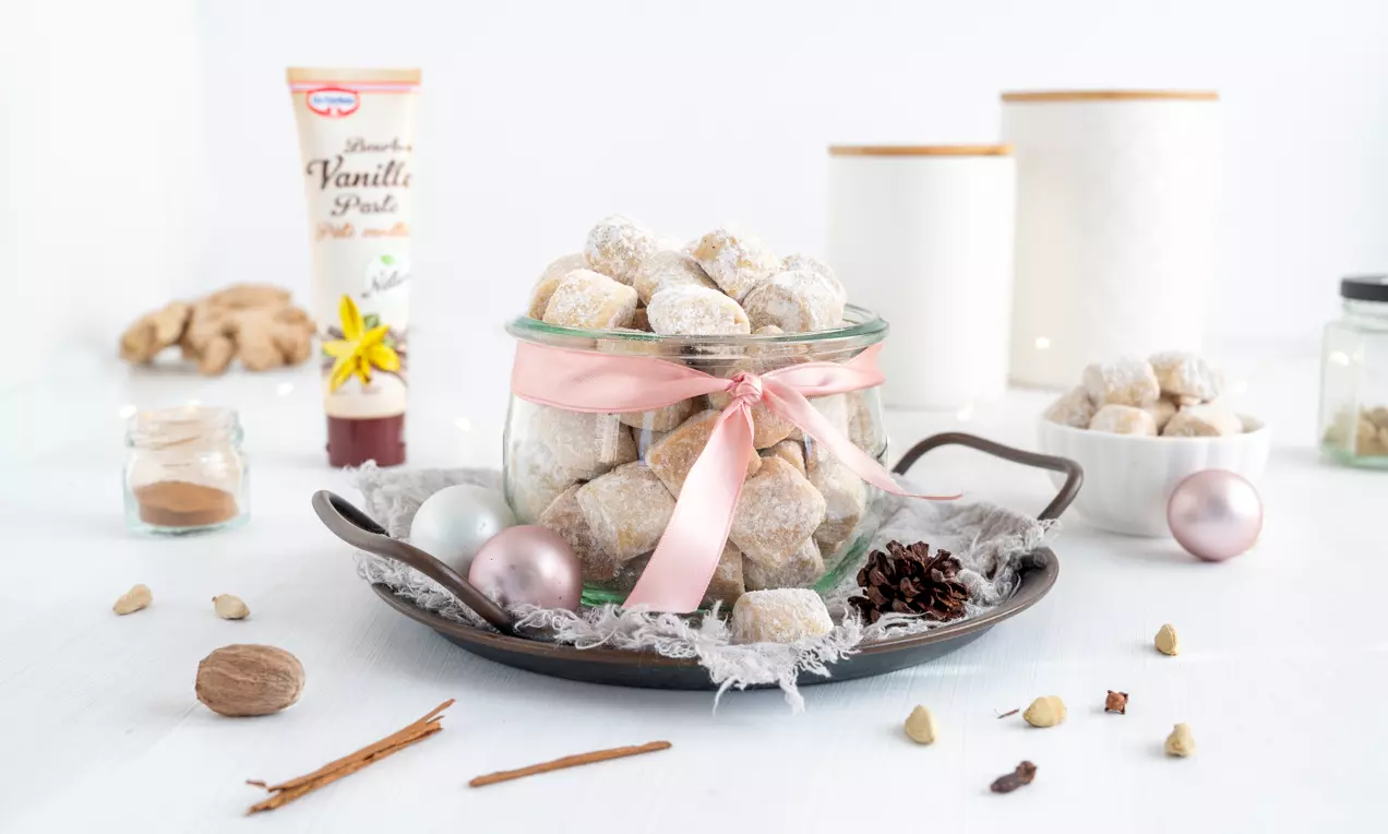 Chai Traumstücke, hellbeige und in Puderzucker gewälzt, in einem Glas mit rosa Schleife auf einem Tablett mit Tannenzapfen und Deko drapiert